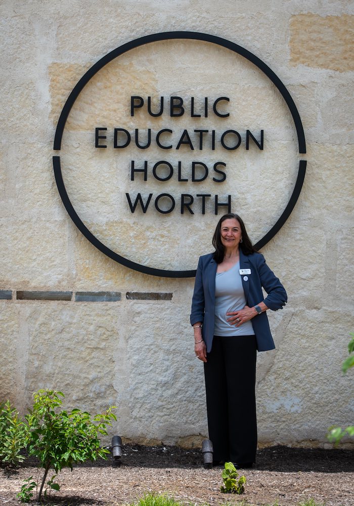 Pauline Dow, vice president of The Holdsworth Center, is photographed posing beneath a metal badge mounted on the administration building on the Campus at Lake Austin.