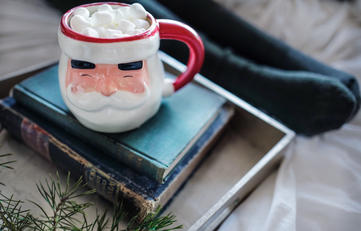 A hot chocolate and marshmallow filled santa mug sits atop a pile of books.