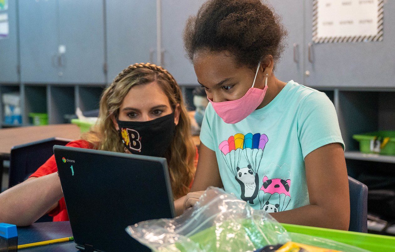 A teacher and student, both wearing masks, are photographed working together in front of a laptop computer in class.