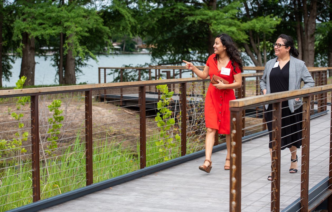 Amy O'Connor, Teacher at Kaiser Elementary in Klein ISD, and Betty Zavala, Principal at Kaiser Elementary in Klein ISD, are photographed smiling as they walk along a bridge at the Campus and Lake Austin during a Holdsworth Center Campus Leadership Program session.
